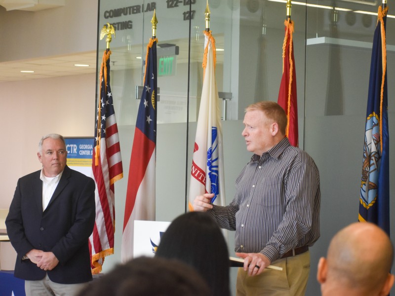 Two men standing. One of them is speaking behind a podium. The other man is standing next to him.