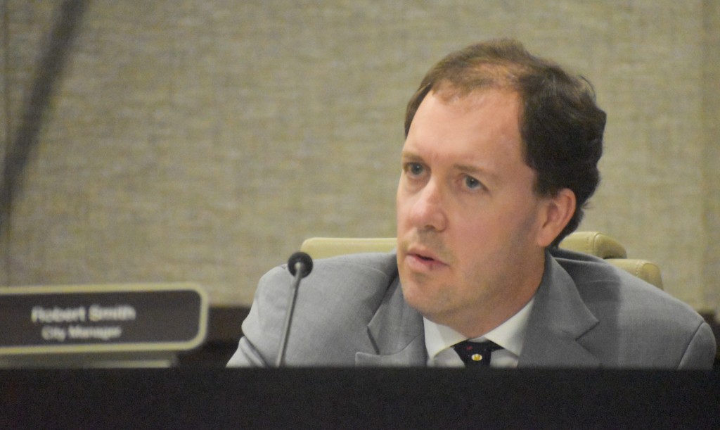 A man in a gray suit sitting behind a desk. He is speaking into a microphone.