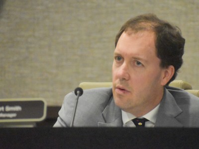 A man in a gray suit sitting behind a desk. He is speaking into a microphone.