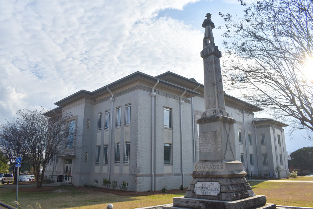 A tall gray statue standing in front of a large gray building. There is a Confederate soldier standing on top of the monument.  It is a sunny day. 