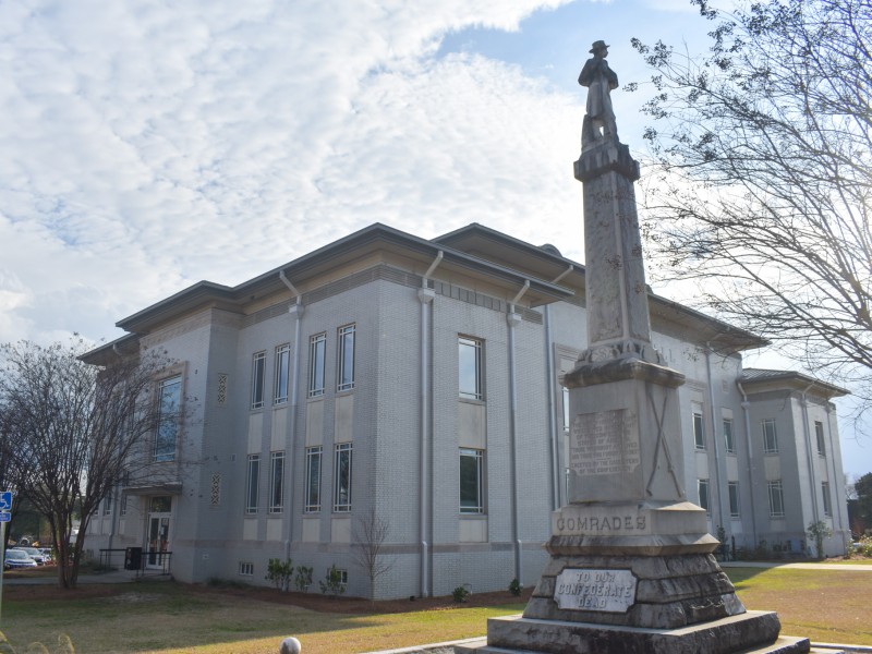 A tall gray statue standing in front of a large gray building. It is a sunny day.