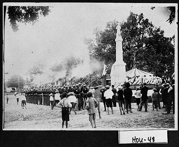 An old photograph of the monument, which is a tall gray statue with a Confederate soldier standing at the top. There are people surrounding the statue and there is smoke in the air. 