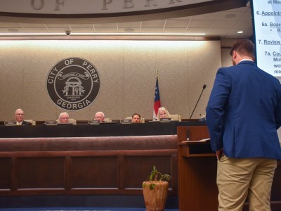 A man is standing behind a podium on the right. He is facing a table with men and women sitting behind it.
