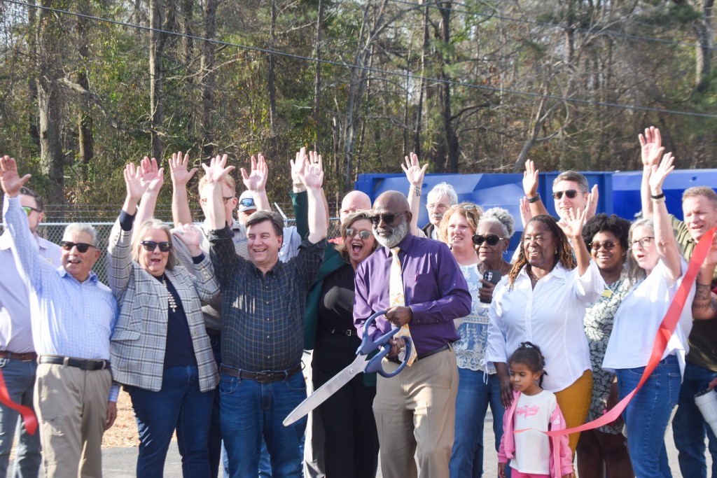 A group of people smiling and their hands are in the air. A man wearing a purple shirt stands in the middle holding large scissors. He is smiling. 