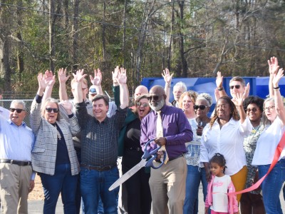 A group of people smiling and their hands are in the air. A man wearing a purple shirt stands in the middle holding large scissors. He is smiling.