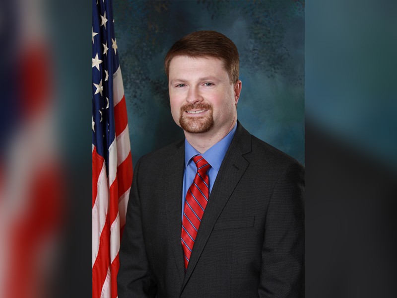 A man smiling. He is wearing a suit and a red tie.