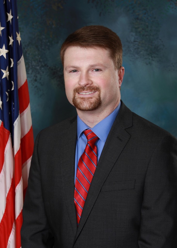 A man smiling. He is wearing a suit and a red tie.