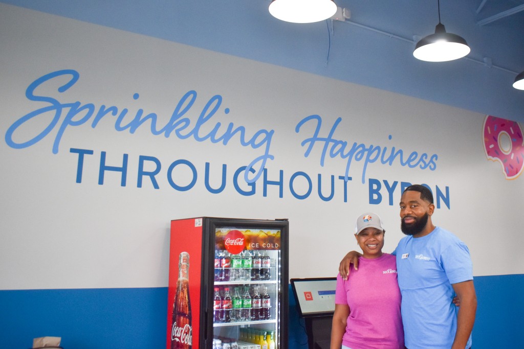 A man and woman smiling. The woman is wearing a pink shirt and the man is wearing a blue shirt. Behind them is a sign written in blue lettering. It says: "Sprinkling happiness throughout Byron." 
