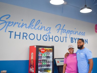 A man and woman smiling. The woman is wearing a pink shirt and the man is wearing a blue shirt. Behind them is a sign written in blue lettering. It says: "Sprinkling happiness throughout Byron."