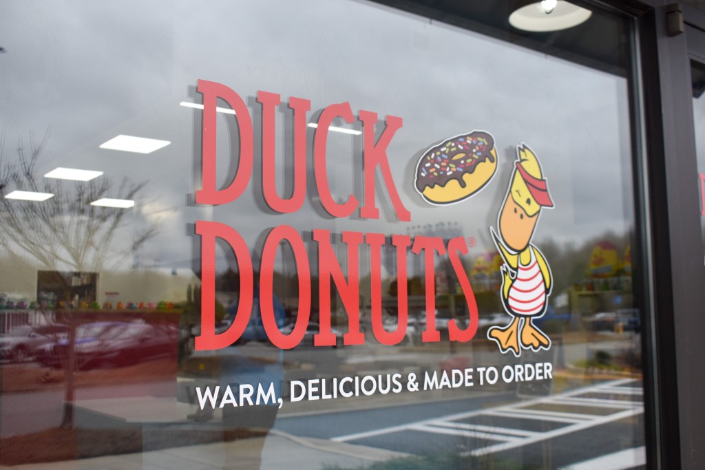 A sign outside a door. It is written in red lettering that says, "Duck Donuts." There is a yellow duck and a chocolate donut painted next to the words.