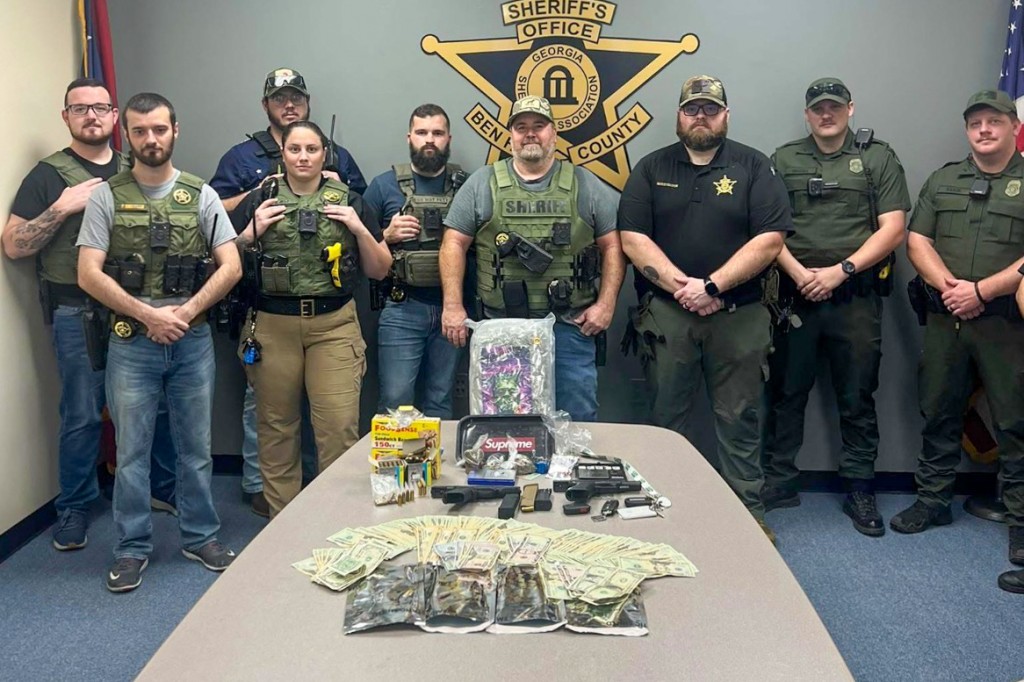 Men in uniform standing behind a table. The table has the evidence including money, firearms and bags of marijuana.