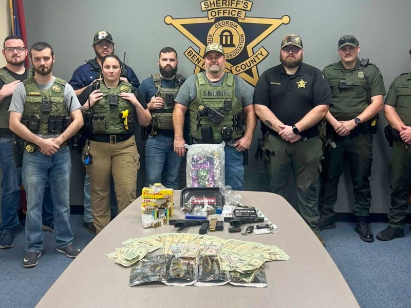 Men in uniform standing behind a table. The table has the evidence including money, firearms and bags of marijuana.