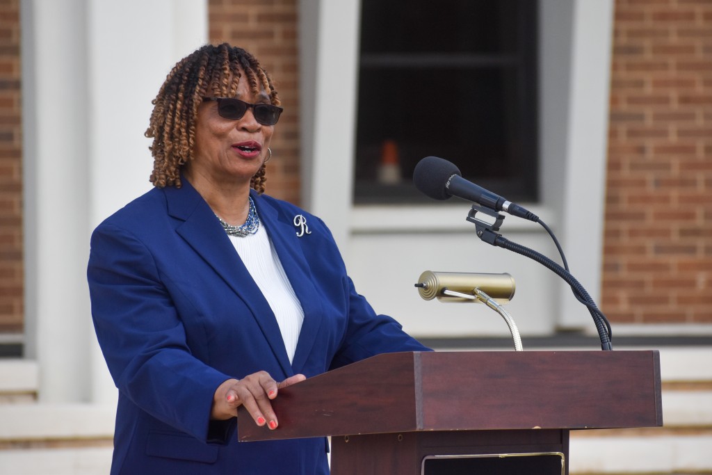 A woman in a blue suit speaking behind a brown podium. She is wearing sunglasses. 