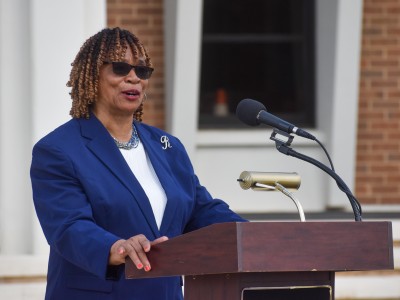 A woman in a blue suit speaking behind a brown podium. She is wearing sunglasses.
