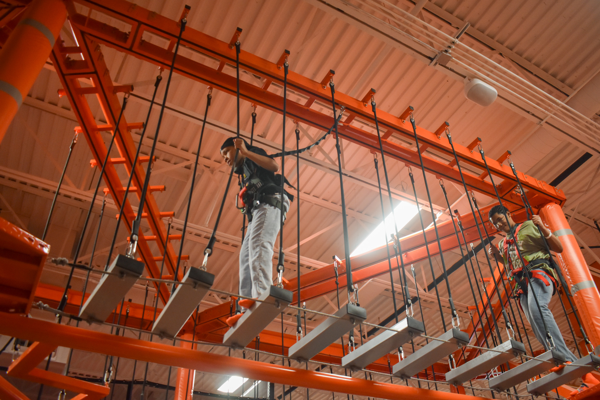 Two boys walking across an obstacle in the air. They are being held by ropes. 