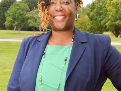 A women in a blue blazer and green shirt smiling. She has one hand on her hip.
