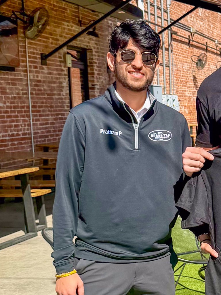 A man in a black zip-up shirt smiling. He is wearing sunglasses. 