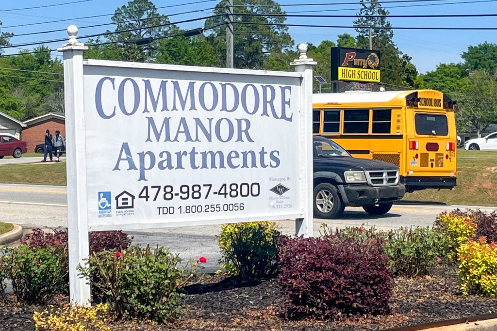 A white sign with blue lettering. In the background there is school bus passing by. 