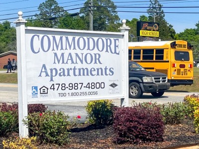 A white sign with blue lettering. In the background there is school bus passing by.