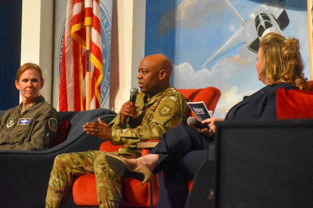 A man sitting in the middle wearing a military uniform. He is speaking into a microphone. Two women sit on his left and right, listening to him. 
