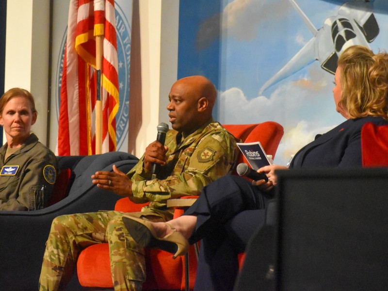A man sitting in the middle wearing a military uniform. He is speaking into a microphone. Two women sit on his left and right, listening to him.