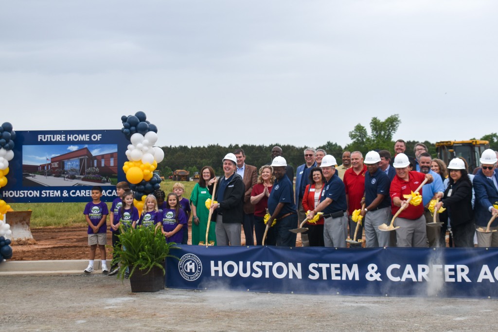 A group of people wearing hard hats and shovels. Some are smiling. Next to them are a group of children, also smiling.