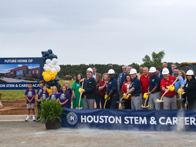 A group of people wearing hard hats and shovels. Some are smiling. Next to them are a group of children, also smiling.