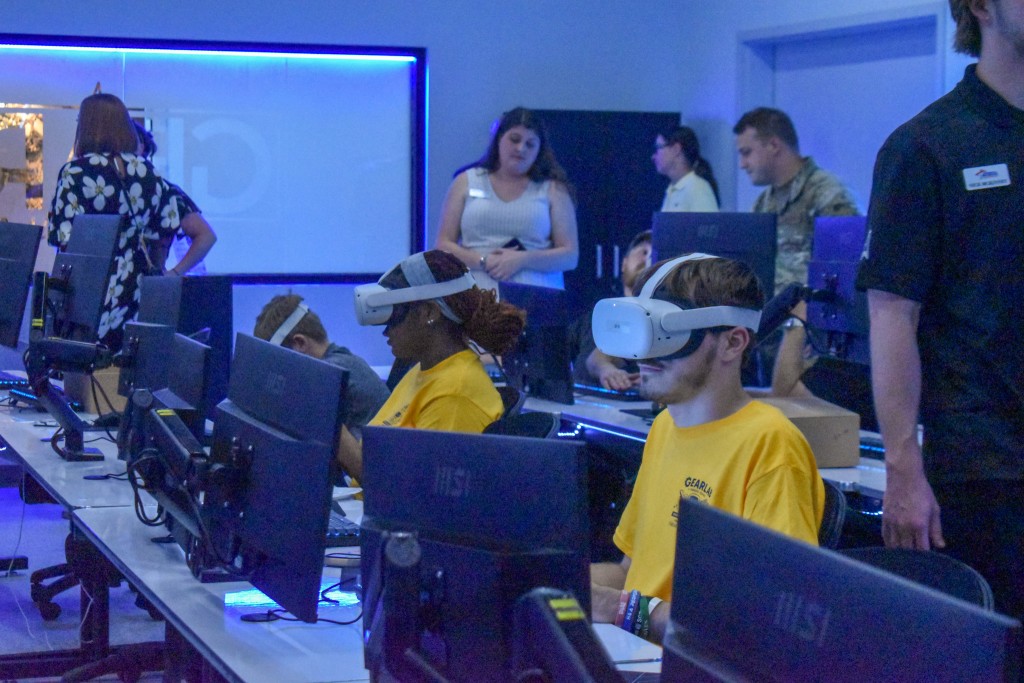 A boy and girl sitting behind a computer. They are wearing virtual reality goggles.