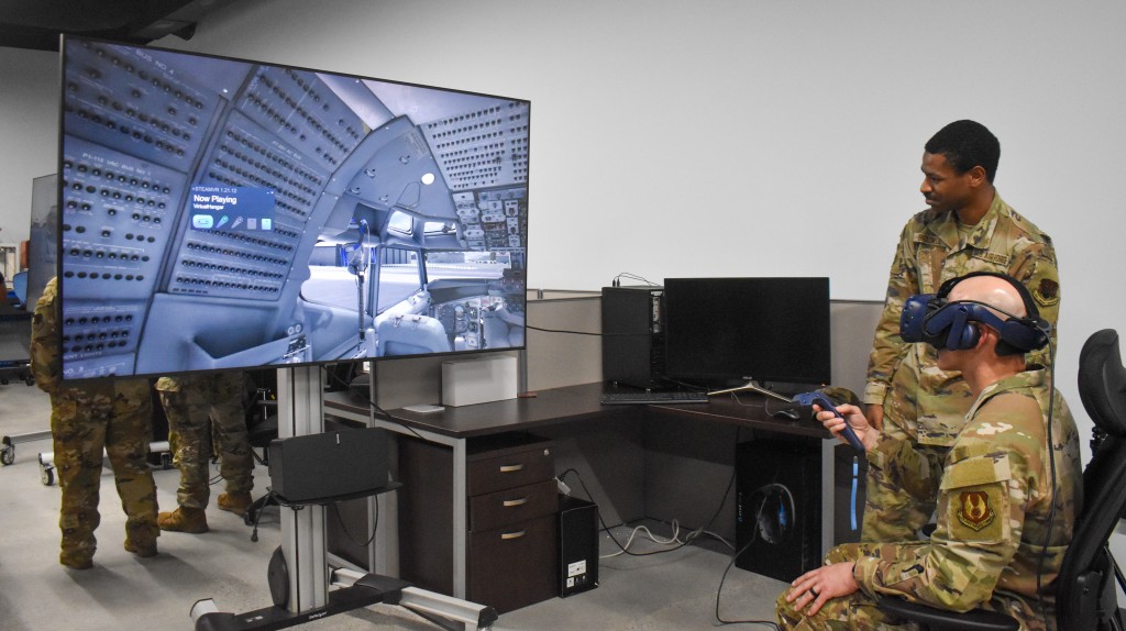 Two men in military uniform. One of them is sitting in front of a TV and wearing virtual reality goggles while the other is standing beside him.