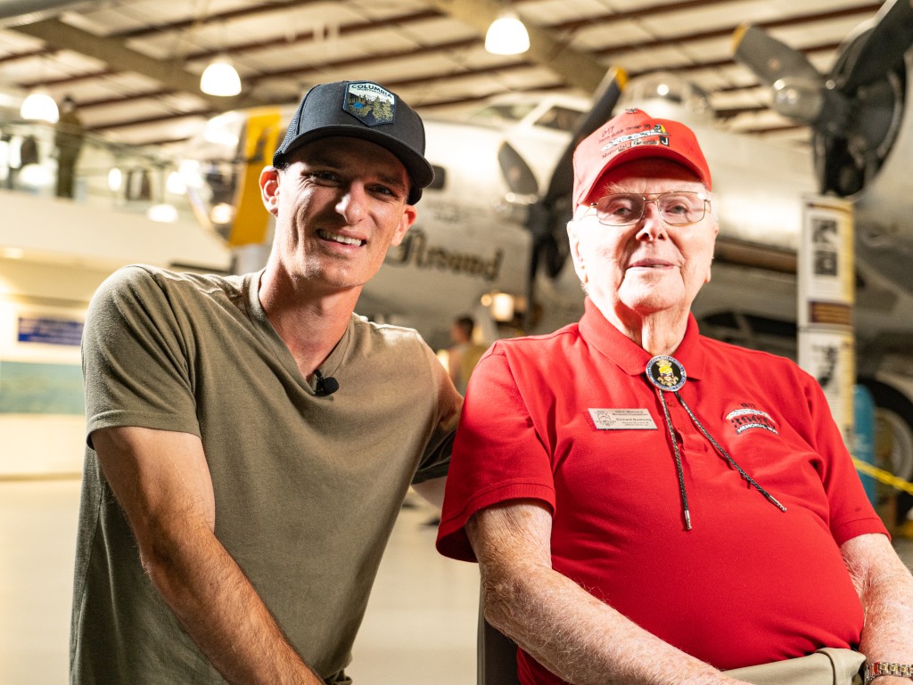 Two men smiling. The one on the left is wearing a hat and gray shirt. The other man is older, wearing a red hat and red shirt. 