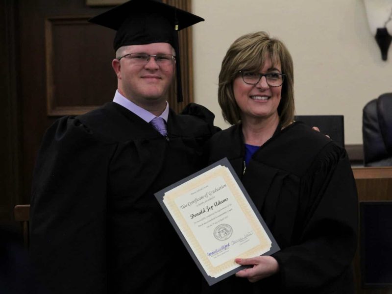 White male in graduate out fit with a female judge. The judge is holding a graduation certificate.