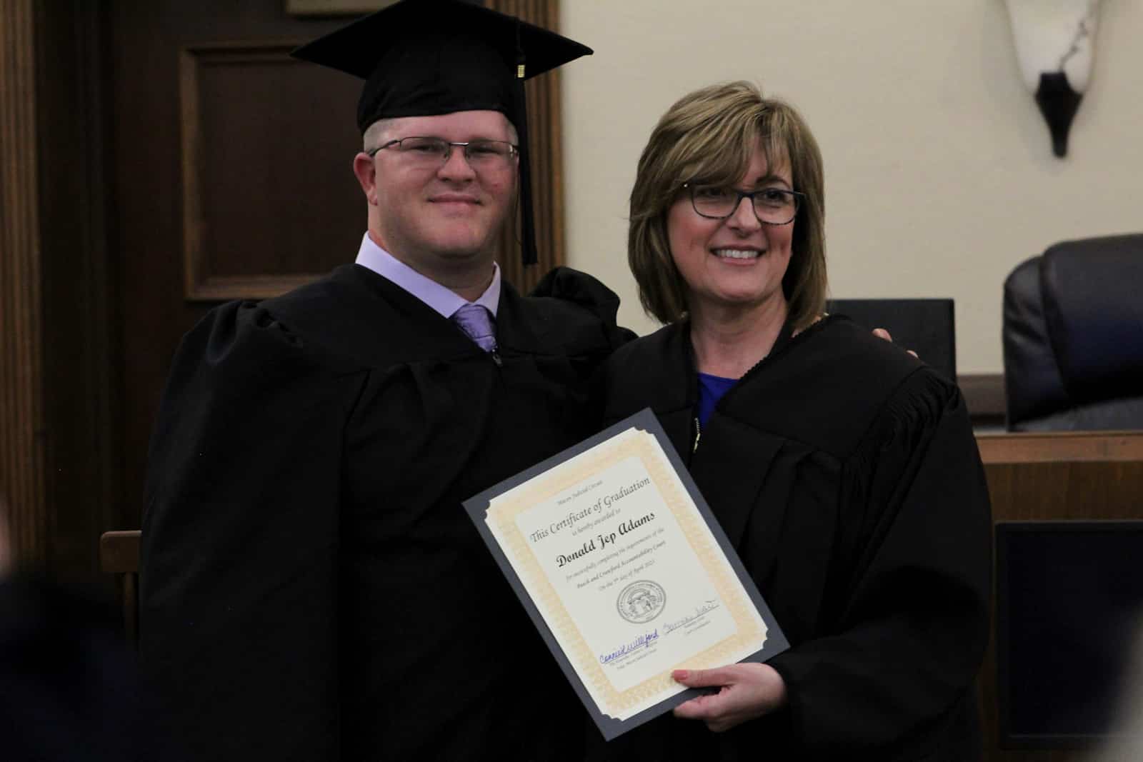 White male in graduate out fit with a female judge. The judge is holding a graduation certificate.