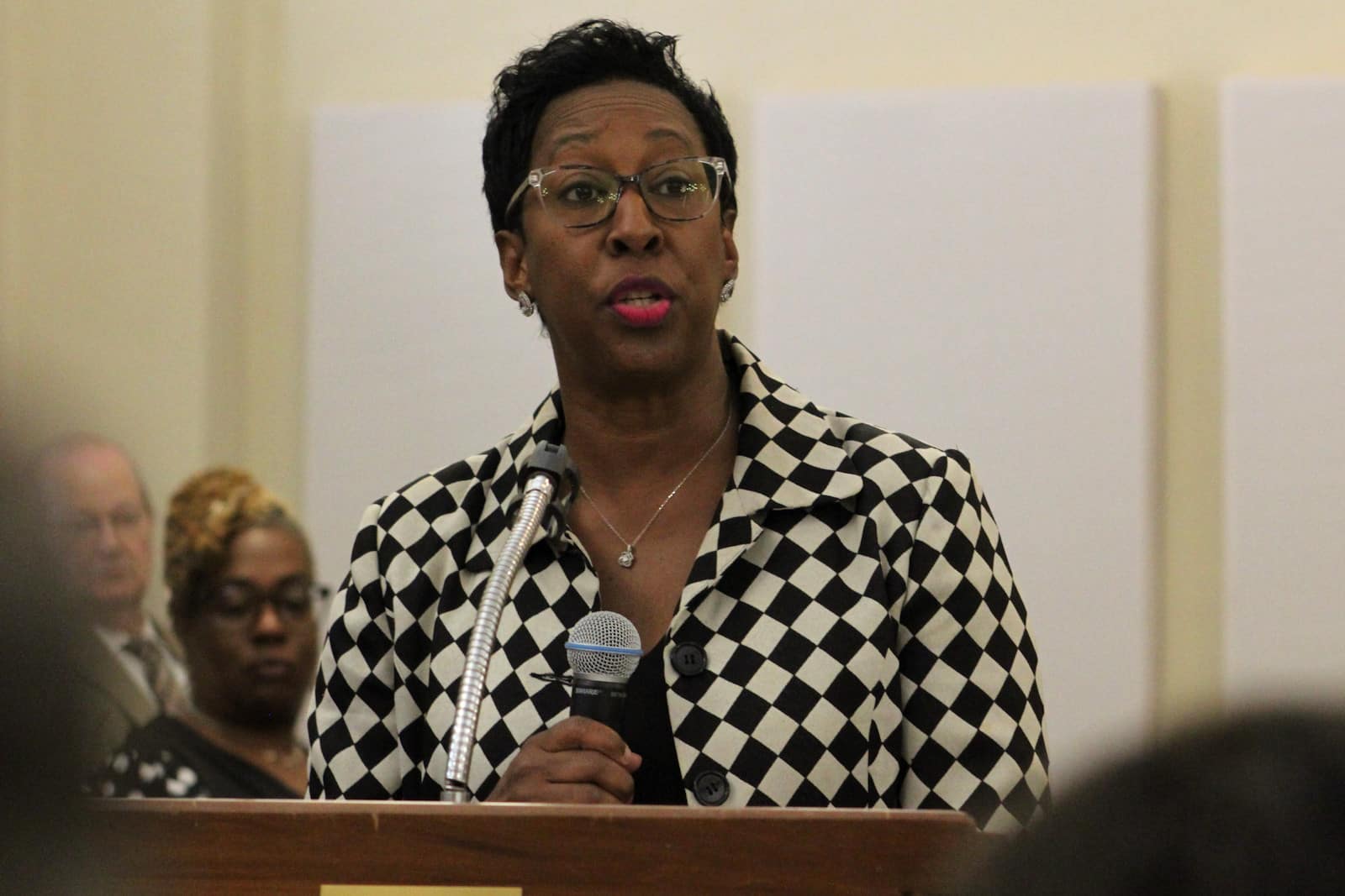 African American Female with shorter hair and clear glasses, speaks at a podium while holding a microphone