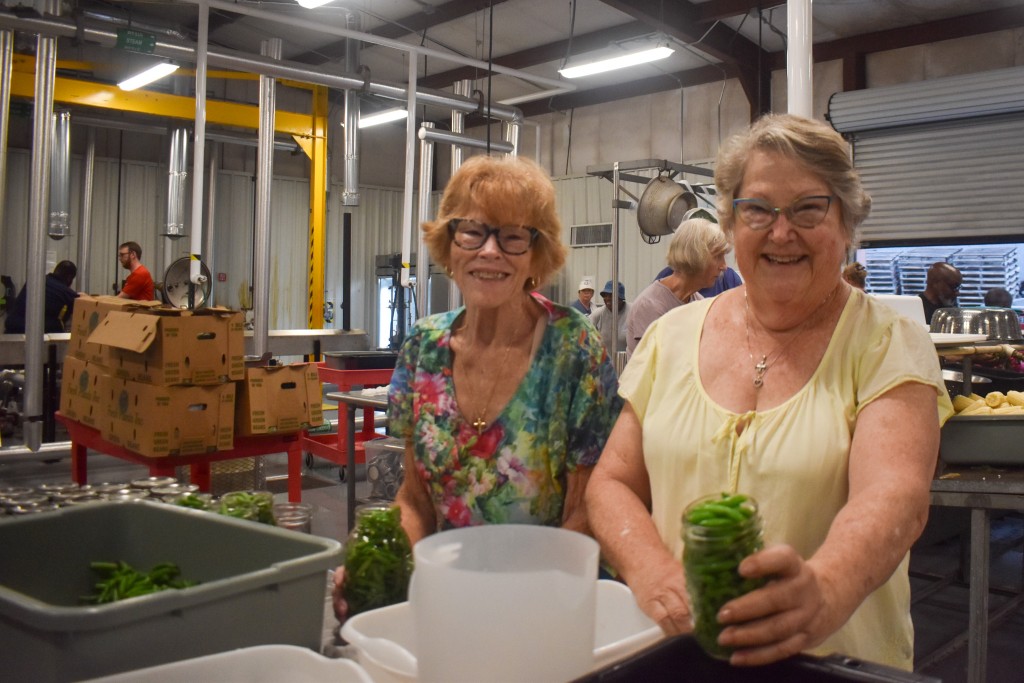 Two older women smiling. The woman on the right is holding a jar of green beans.