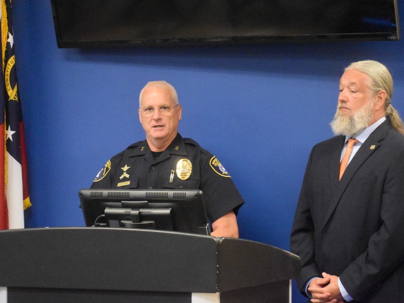 One man in a uniform speaking behind a podium. A man in a black suit is standing next to him.