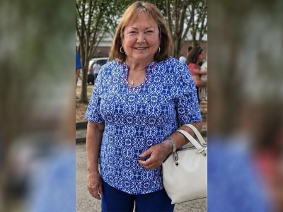 A white woman in a blue shirt. She has brown hair and is smiling. The photo is against a blurry background.