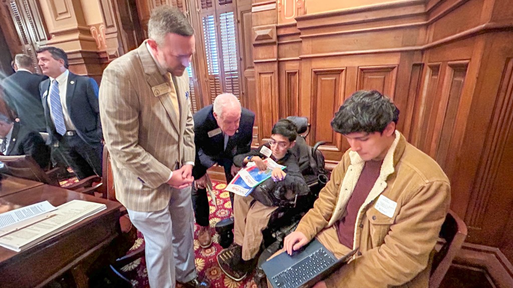 Four men talking together. Two of the men are older wearing suits. The other two are sitting down. One of them is holding a computer showing one of the older man their screen. 