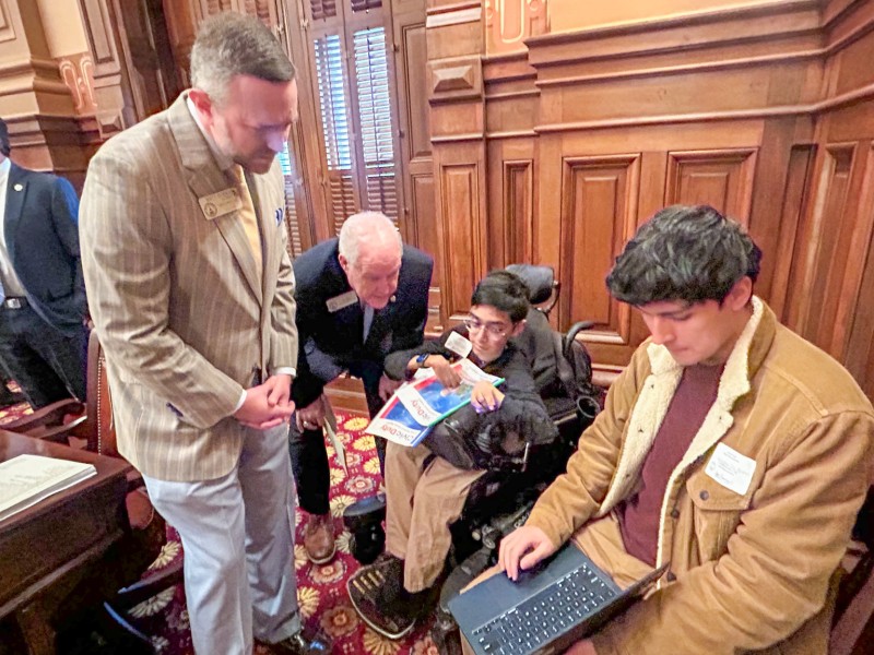Four men talking together. Two of the men are older wearing suits. The other two are sitting down. One of them is holding a computer showing one of the older man their screen.