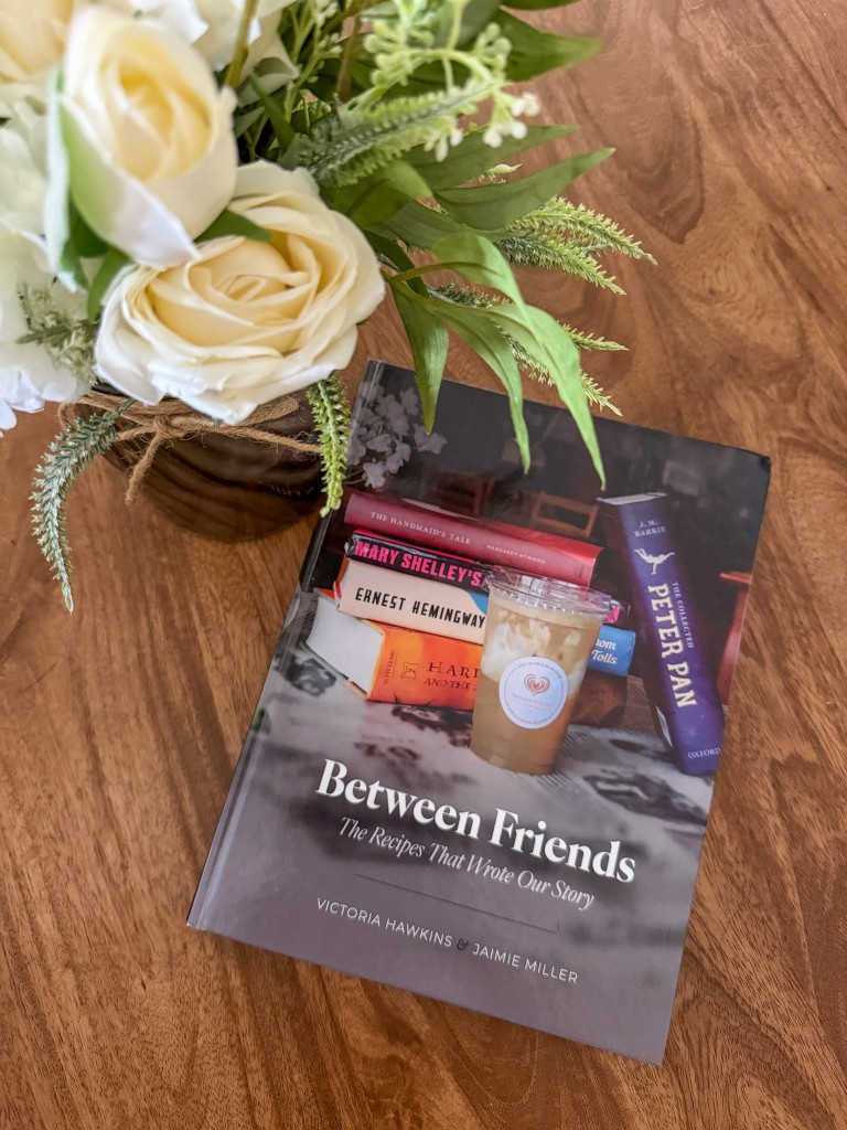 A book on a wooden table next to white flowers. The cover is a stack of books and a cup of iced coffee next to it. 