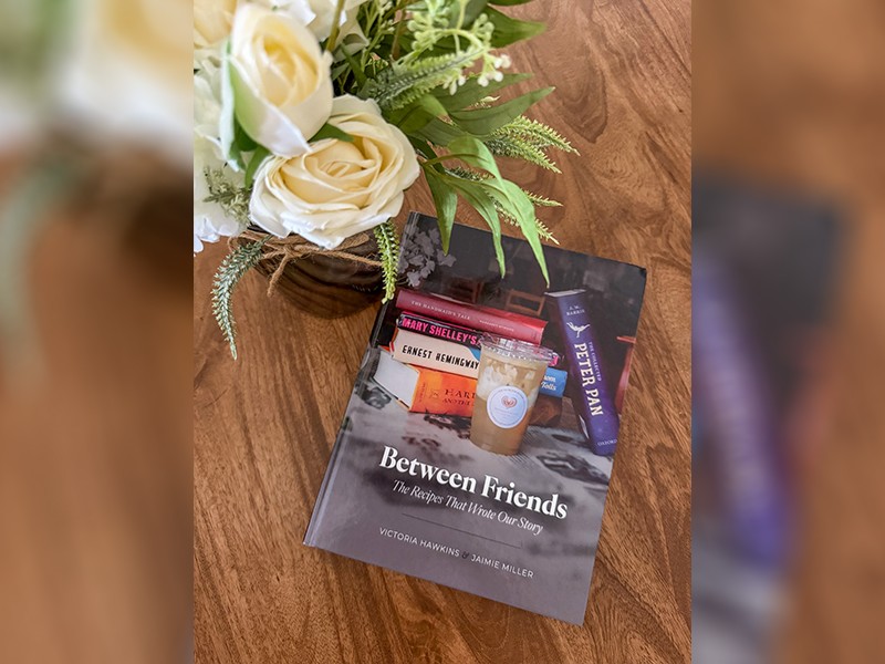 A book on a wooden table next to white flowers. The cover is a stack of books and a cup of iced coffee next to it.