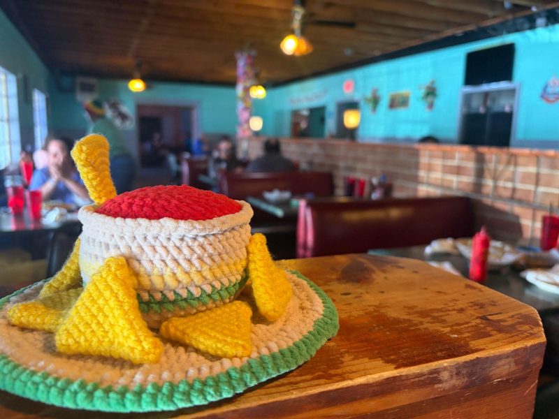 A knitted bowl made to look like chips and salsa sits on a wooden partition, colorful decor in the background.