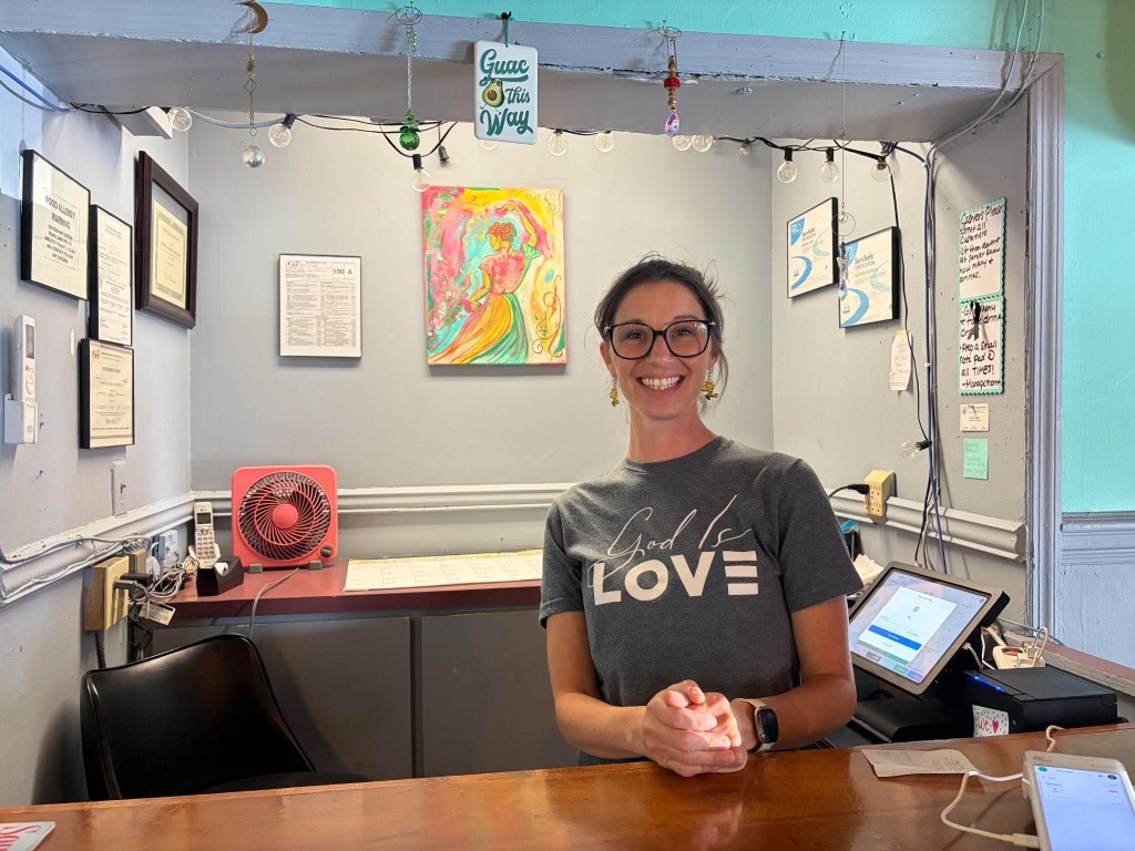 A smiling young woman wearing glasses behind the front counter of a restaurant.