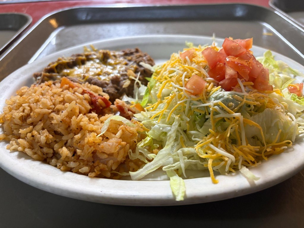 A plate with a half chalupa, rice and beans. The chalupa is topped with shredded cheese, ground beef, lettuce, tomato, and guacamole.