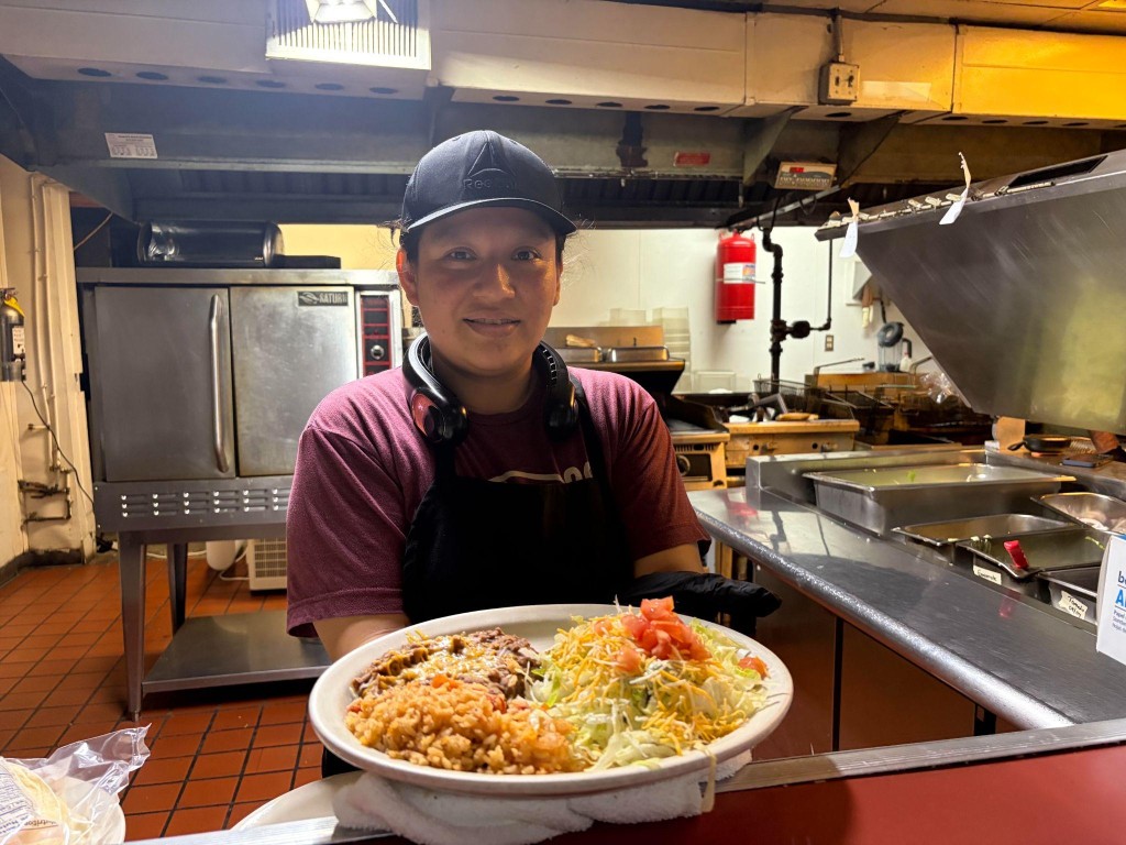 A young woman holds a plate of chalupas, rice and beans in a commercial kitchen.