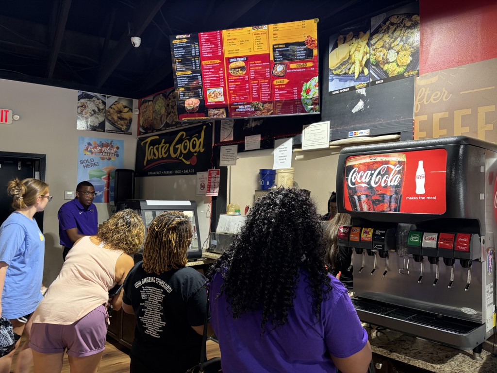 Customers line up at counter of Taste Good Restaurant with the menu board overhead, a Coca-Cola drink dispenser on the right and the restaurant logo in the background. The photo gives an idea of how small the space is for the restaurant in the corner of the convenience store.