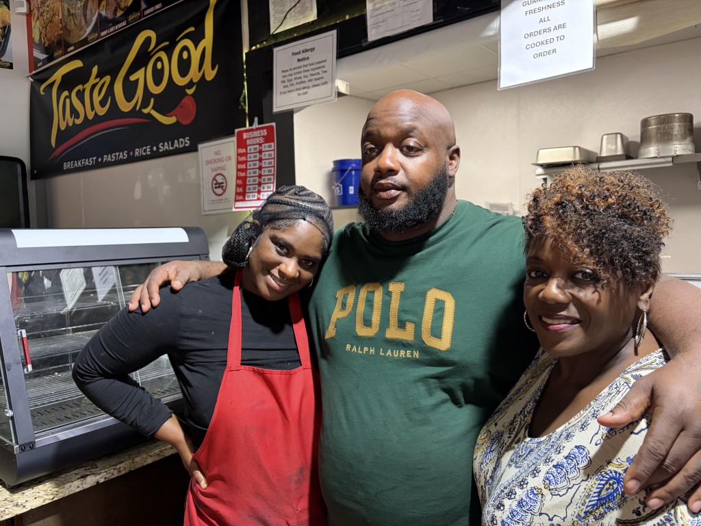 Taste Good Restaurant owner with his arms around his mom and girlfriend inside the takeout spot with the restaurant logo shown n the background.