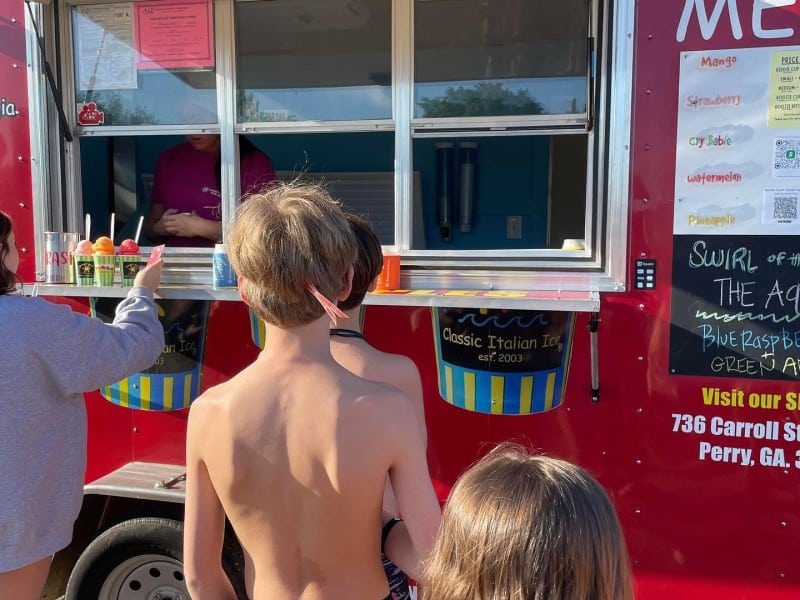 Photo is taken from behind kids lined up at the mobile unit of Sunset Slush of Middle Georgia. The vehicle is read and has a window from which to order and receive the colorful Italian ice in cups.