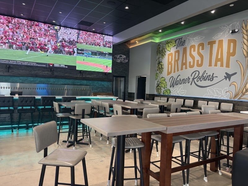 A super large TV screen shows a football game and a baseball game at the back of the craft beer bar above the 60-plus taps with a large L shaped bar and high tables with high seats. The Brass Tap Warner Robins is painted on a side wall with an airplane flying out of the R in Robins.