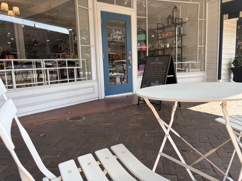 Closeup of storefront shows small white table with foldable white chair with storefront in the background. The storefront has large glass windows and a door with a long window inside of blue/green trim.