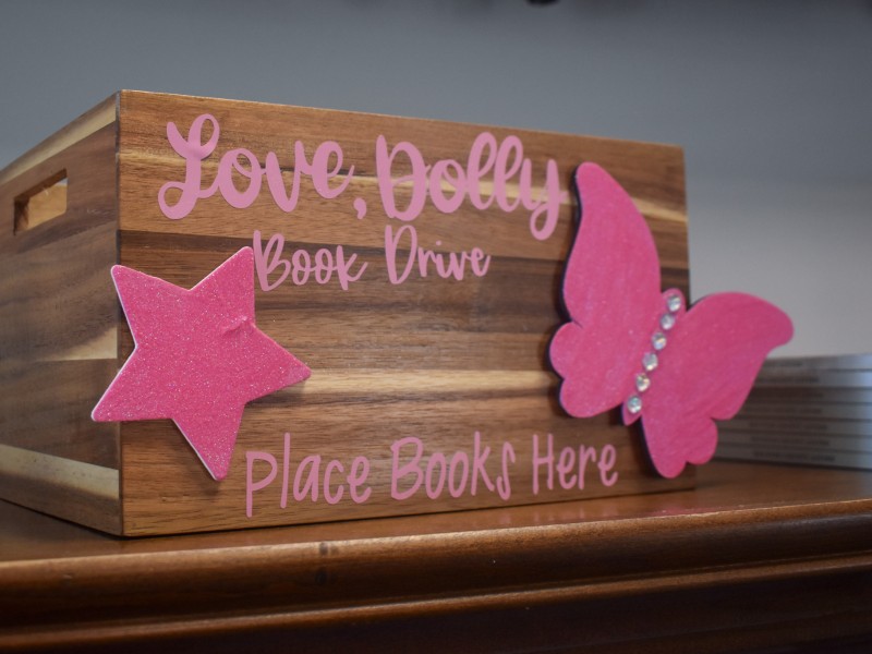 A brown crate with pink lettering saying, "Love, Dolly book drive. Place book here." It is decorated with a pink star and butterfly.